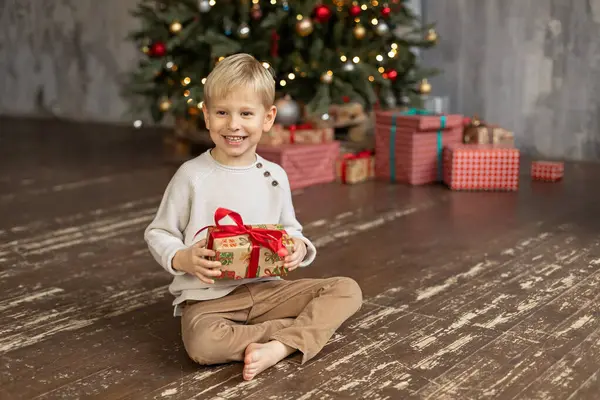 Küçük sarışın bir çocuk ahşap bir zeminde oturuyor, elinde bir Noel hediyesi tutuyor ve gülüyor. Arka planda hediyeli bir Noel ağacı var..