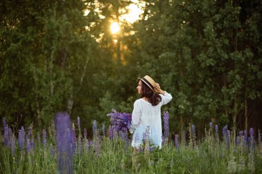 Gün batımında tarlada bir buket lupinle hasır şapkalı güzel bir genç kadın.
