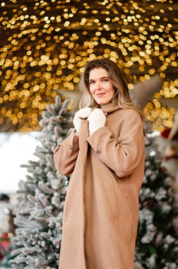 Palto ve beyaz eldivenli mutlu kadın Noel çelenklerinin önünde duruyor, bayram havasında.