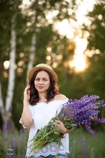 Gün batımında tarlada bir buket lupinle hasır şapkalı güzel bir genç kadın.