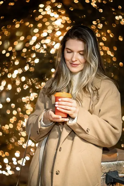 Bej ceketli güzel bir genç kız. Elinde bir bardak kahveyle arka planda bir sürü bokeh ışığı var.