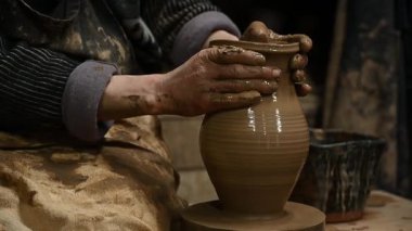 Çömlekçi ellerinin, dönen çömlek çarkında kil çömleği şekillendirdiği yakın plan. Islak kille kaplı sanatçının elleri, geleneksel seramiğin işçiliğini ve dokunsal doğasını ön plana çıkarıyor. Kırsal atölye ayarları gerçeklik katıyor
