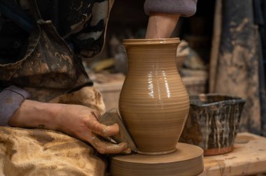 Çömlek çarkında seramik vazoyu dikkatlice şekillendirirken ıslak çamura bulanmış çömlekçi ellerini kapatın. Zanaatkar, kenarı rafine etmek için ahşap bir alet kullanıyor, geleneksel çömlekçiliğin işçiliğini ve dokunsal doğasını sergiliyor.. 