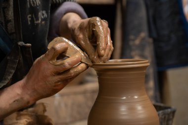 Çömlek çarkında seramik vazoyu dikkatlice şekillendirirken ıslak çamura bulanmış çömlekçi ellerini kapatın. Zanaatkar, kenarı rafine etmek için ahşap bir alet kullanıyor, geleneksel çömlekçiliğin işçiliğini ve dokunsal doğasını sergiliyor.. 