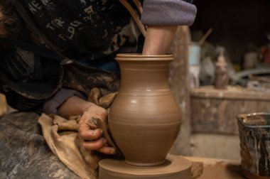 Çömlek çarkında seramik vazoyu dikkatlice şekillendirirken ıslak çamura bulanmış çömlekçi ellerini kapatın. Zanaatkar, kenarı rafine etmek için ahşap bir alet kullanıyor, geleneksel çömlekçiliğin işçiliğini ve dokunsal doğasını sergiliyor.. 