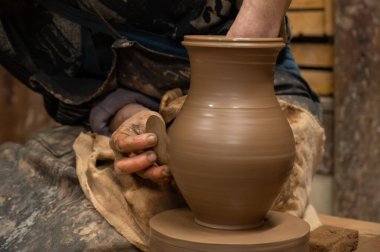 Çömlek çarkında seramik vazoyu dikkatlice şekillendirirken ıslak çamura bulanmış çömlekçi ellerini kapatın. Zanaatkar, kenarı rafine etmek için ahşap bir alet kullanıyor, geleneksel çömlekçiliğin işçiliğini ve dokunsal doğasını sergiliyor.. 