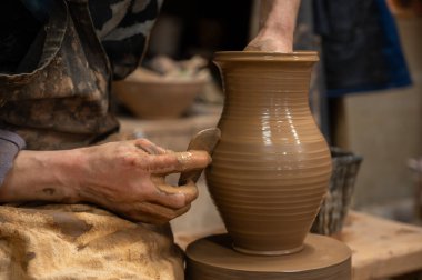Çömlek çarkında seramik vazoyu dikkatlice şekillendirirken ıslak çamura bulanmış çömlekçi ellerini kapatın. Zanaatkar, kenarı rafine etmek için ahşap bir alet kullanıyor, geleneksel çömlekçiliğin işçiliğini ve dokunsal doğasını sergiliyor.. 