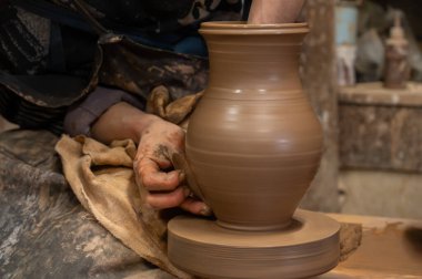 Çömlek çarkında seramik vazoyu dikkatlice şekillendirirken ıslak çamura bulanmış çömlekçi ellerini kapatın. Zanaatkar, kenarı rafine etmek için ahşap bir alet kullanıyor, geleneksel çömlekçiliğin işçiliğini ve dokunsal doğasını sergiliyor.. 