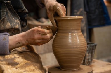 Çömlek çarkında seramik vazoyu dikkatlice şekillendirirken ıslak çamura bulanmış çömlekçi ellerini kapatın. Zanaatkar, kenarı rafine etmek için ahşap bir alet kullanıyor. Geleneksel çömlekçiliğin işçiliği ve dokunsal doğasını sergiliyor.. 
