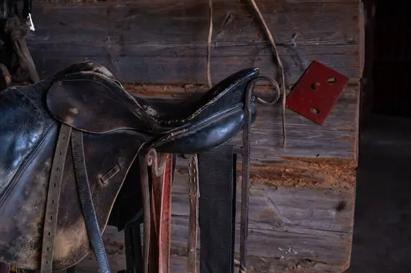 Eski siyah deri bir eyer görünür aşınma ve yırtık taşralı ahşap bir ahırda metal bir standın üzerinde duruyor. Eyerin eski dokusu ve arka plandaki pürüzlü ahşap duvar nostaljik ve otantik bir binicilik atmosferi yaratıyor.