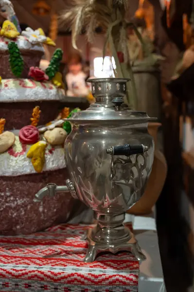 Geleneksel işlemeli bir kumaş üzerinde klasik bir metal semaver, arka planda dekoratif şenlik ekmeği olan kırsal bir iç mekan..