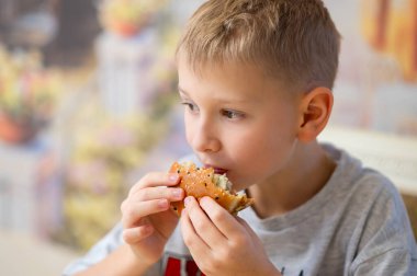 Kısa sarı saçlı genç bir çocuk lezzetli bir burgerin tadını çıkarıyor, büyük bir ısırık alıyor. Susamlı ekmeği iki eliyle tutuyor, tadını çıkarıyor. Bulanık arka plan sahneye sıcaklık ve samimiyet ekler..