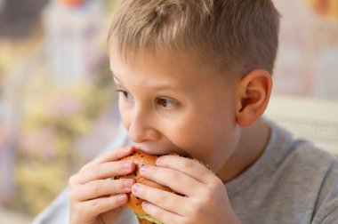 Kısa sarı saçlı genç bir çocuk lezzetli bir burgerin tadını çıkarıyor, büyük bir ısırık alıyor. Susamlı ekmeği iki eliyle tutuyor, tadını çıkarıyor. Bulanık arka plan sahneye sıcaklık ve samimiyet ekler..