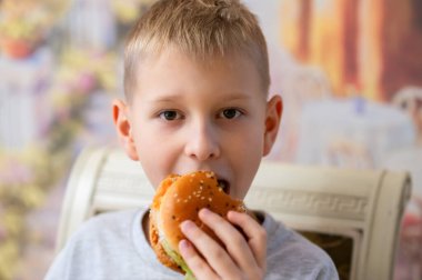 Kısa sarı saçlı mutlu genç bir çocuk lezzetli bir burgerin tadını çıkarıyor, zevkle gülümsüyor. Kısmen yenmiş sandviçi iki elinde tutuyor. Bulanık arka plan rahat ve sıcak bir atmosfer yaratır..