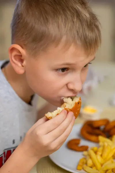 Genç bir çocuk yemeğini yerken, hamburgerinden bir ısırık alıyor. Tabağı patates kızartması ve soğan halkalarıyla dolu. Yakın çekim yüz ifadesini ve lezzetli fast-food setini yakalıyor..
