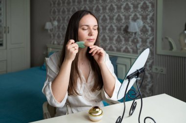 İpek bornozlu genç bir kadın rahat bir yatak odasında gösterişli bir aynada otururken yeşim bir gua sha yüz aracı kullanıyor. Güzelliği ve cilt bakımını takip eder.