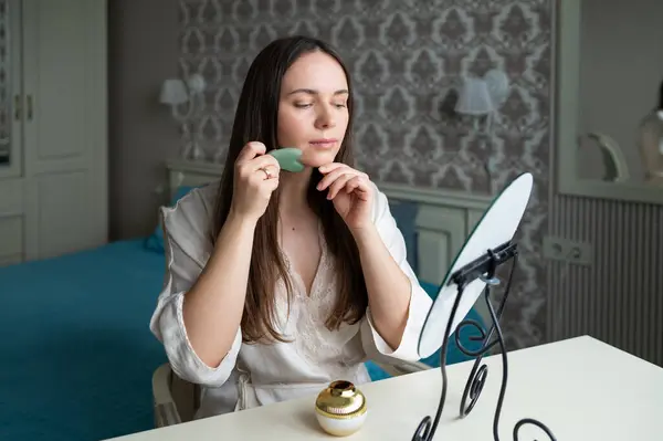 İpek bornozlu genç bir kadın rahat bir yatak odasında gösterişli bir aynada otururken yeşim bir gua sha yüz aracı kullanıyor. Güzelliği ve cilt bakımını takip eder.