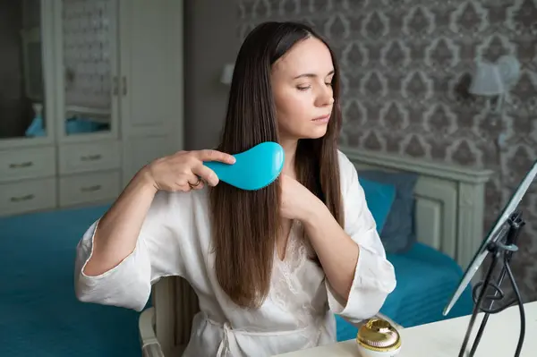 İpek bornozlu genç bir kadın, rahat bir yatak odasında gösterişli bir aynanın karşısında otururken uzun düz saçlarını mavi bir tarakla tarıyor. Sabah güzellik rutini
