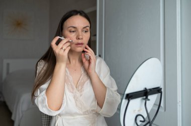 İpek bornozlu orta yaşlı bir kadın aynaya bakarken kendine endişeli bir ifadeyle güzellik aşısı yapıyor. Evde güzellik ve cilt bakımı