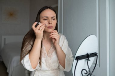 İpek bornozlu orta yaşlı bir kadın aynaya bakarken kendine endişeli bir ifadeyle güzellik aşısı yapıyor. Evde güzellik ve cilt bakımı