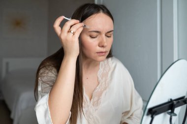 İpek bornozlu orta yaşlı bir kadın aynaya bakarken kendine endişeli bir ifadeyle güzellik aşısı yapıyor. Evde güzellik ve cilt bakımı