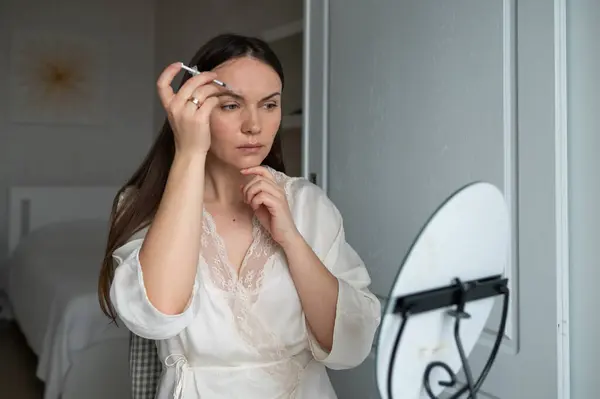 İpek bornozlu orta yaşlı bir kadın aynaya bakarken kendine endişeli bir ifadeyle güzellik aşısı yapıyor. Evde güzellik ve cilt bakımı