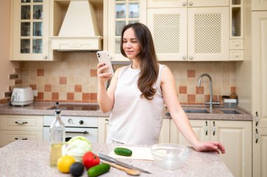 Mutfakta taze sebze salatası hazırlarken akıllı telefon kullanan gülümseyen kadın. Sağlıklı yaşam tarzı, ev yemekleri ve online tarif ilhamı.
