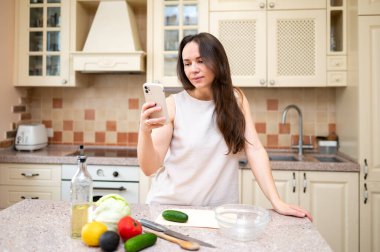 Mutfakta taze sebze salatası hazırlarken akıllı telefon kullanan gülümseyen kadın. Sağlıklı yaşam tarzı, ev yemekleri ve online tarif ilhamı.