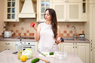Parlak mutfakta elinde olgun domates ve avokadoyla salata hazırlamaya giden genç bir kadın. Taze sebzeler ve kesim tahtası masada.