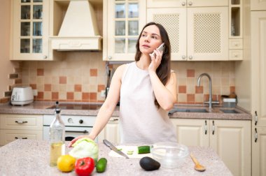 Parlak mutfakta telefonla konuşan, gülümseyen ve salata yapmayı planlayan genç bir kadın. Taze sebzeler ve yemek hazır.