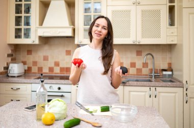 Parlak mutfakta elinde olgun domates ve avokadoyla salata hazırlamaya giden genç bir kadın. Taze sebzeler ve kesim tahtası masada.