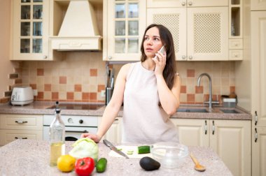 Parlak mutfakta telefonla konuşan, gülümseyen ve salata yapmayı planlayan genç bir kadın. Taze sebzeler ve yemek hazır.