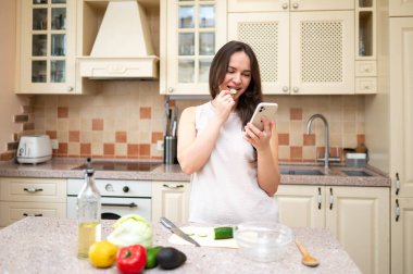 Samimi bir mutfakta genç bir kadın telefonunu kontrol ederken salatalık dilimi yiyor. Tezgahta taze sebzeler var. Sağlıklı yaşam tarzı, ev yemekleri ve dengeli beslenme..