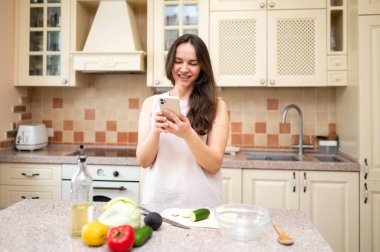 Parlak mutfakta telefonla konuşan, gülümseyen ve salata yapmayı planlayan genç bir kadın. Taze sebzeler ve yemek hazır.