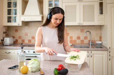 Mutfakta kulaklık takıp sebze doğrayan bir kadın. Taze ürünlerle çevrelenmiş, sakince yemek pişirme işine odaklanmış, müzikten keyif alıyor.