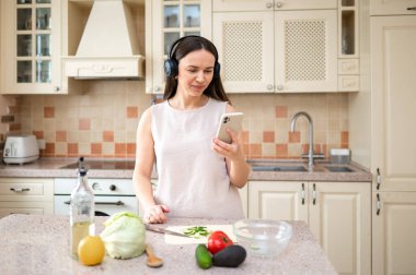  Mutfakta sebze hazırlarken akıllı telefon kullanan bir kadın. Kulaklık takıyor, odaklanmış görünüyor. Tezgahta marul, avokado ve domates gibi taze malzemeler var..