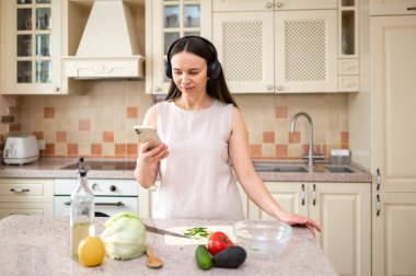  Mutfakta sebze hazırlarken akıllı telefon kullanan bir kadın. Kulaklık takıyor, odaklanmış görünüyor. Tezgahta marul, avokado ve domates gibi taze malzemeler var..