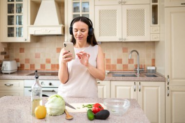  Mutfakta sebze hazırlarken akıllı telefon kullanan bir kadın. Kulaklık takıyor, odaklanmış görünüyor. Tezgahta marul, avokado ve domates gibi taze malzemeler var..