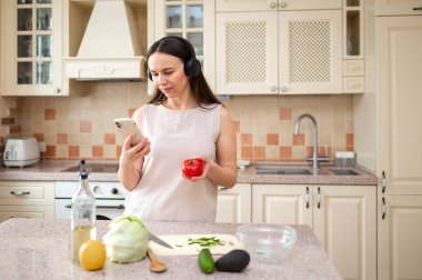 Elinde domates olan bir kadın mutfakta telefonuna bakıyor. Kulaklık takıyor, yemek pişirme ve teknoloji arasında çoklu görev yapıyor, rahat ve odaklanmış bir ifade gösteriyor..