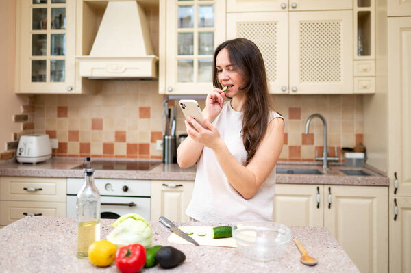 A young woman in a bright kitchen snacks on a cucumber while browsing her phone. Fresh vegetables and ingredients on the counter suggest a healthy and balanced diet