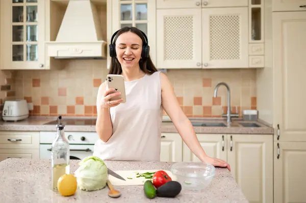  Mutfakta sebze hazırlarken akıllı telefon kullanan bir kadın. Kulaklık takıyor, odaklanmış görünüyor. Tezgahta marul, avokado ve domates gibi taze malzemeler var..