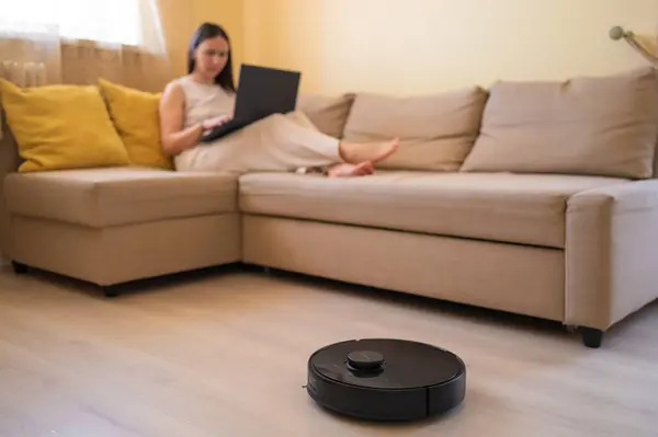 Genç bir kadın rahat bir bej koltukta otururken robotik bir elektrikli süpürge zemini temizliyor. Akıllı ev, otomasyon ve günlük hayatta modern teknoloji. Elektrik süpürgesine odaklan.