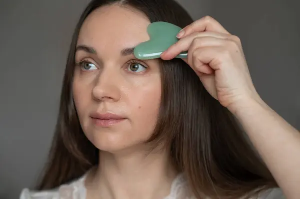 Genç bir kadın yüz masajı için yeşil gua sha kazıyıcı kullanıyor, deri dolaşımını ve rahatlamayı geliştiriyor. Güzellik ve refah için ev cilt bakım rutini.
