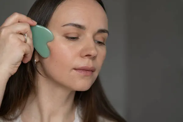 Genç bir kadın yüz masajı için yeşil gua sha kazıyıcı kullanıyor, deri dolaşımını ve rahatlamayı geliştiriyor. Güzellik ve refah için ev cilt bakım rutini.