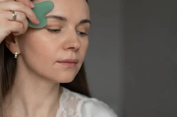Genç bir kadın yüz masajı için yeşil gua sha kazıyıcı kullanıyor, deri dolaşımını ve rahatlamayı geliştiriyor. Güzellik ve refah için ev cilt bakım rutini.