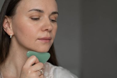 Genç bir kadın yüz masajı için yeşil gua sha kazıyıcı kullanıyor, deri dolaşımını ve rahatlamayı geliştiriyor. Güzellik ve refah için ev cilt bakım rutini.
