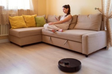 Genç bir kadın rahat bir bej koltukta otururken akıllı telefonunu kullanırken bir robot süpürge yerleri yıkıyor. Akıllı ev, otomasyon ve modern teknolojiler günlük hayatta