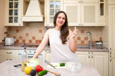 Parlak mutfakta salatalık yiyen ve salata hazırlayan genç bir kadın. Tezgahtaki taze sebzeler ve malzemeler sağlıklı ve dengeli bir diyet öneriyor.
