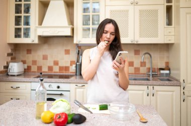 Samimi bir mutfakta genç bir kadın telefonunu kontrol ederken salatalık dilimi yiyor. Tezgahta taze sebzeler var. Sağlıklı yaşam tarzı, ev yemekleri ve dengeli beslenme..