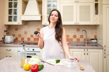 Parlak mutfakta elinde olgun avokadoyla salata hazırlamaya giden genç bir kadın. Taze sebzeler ve kesim tahtası masada.